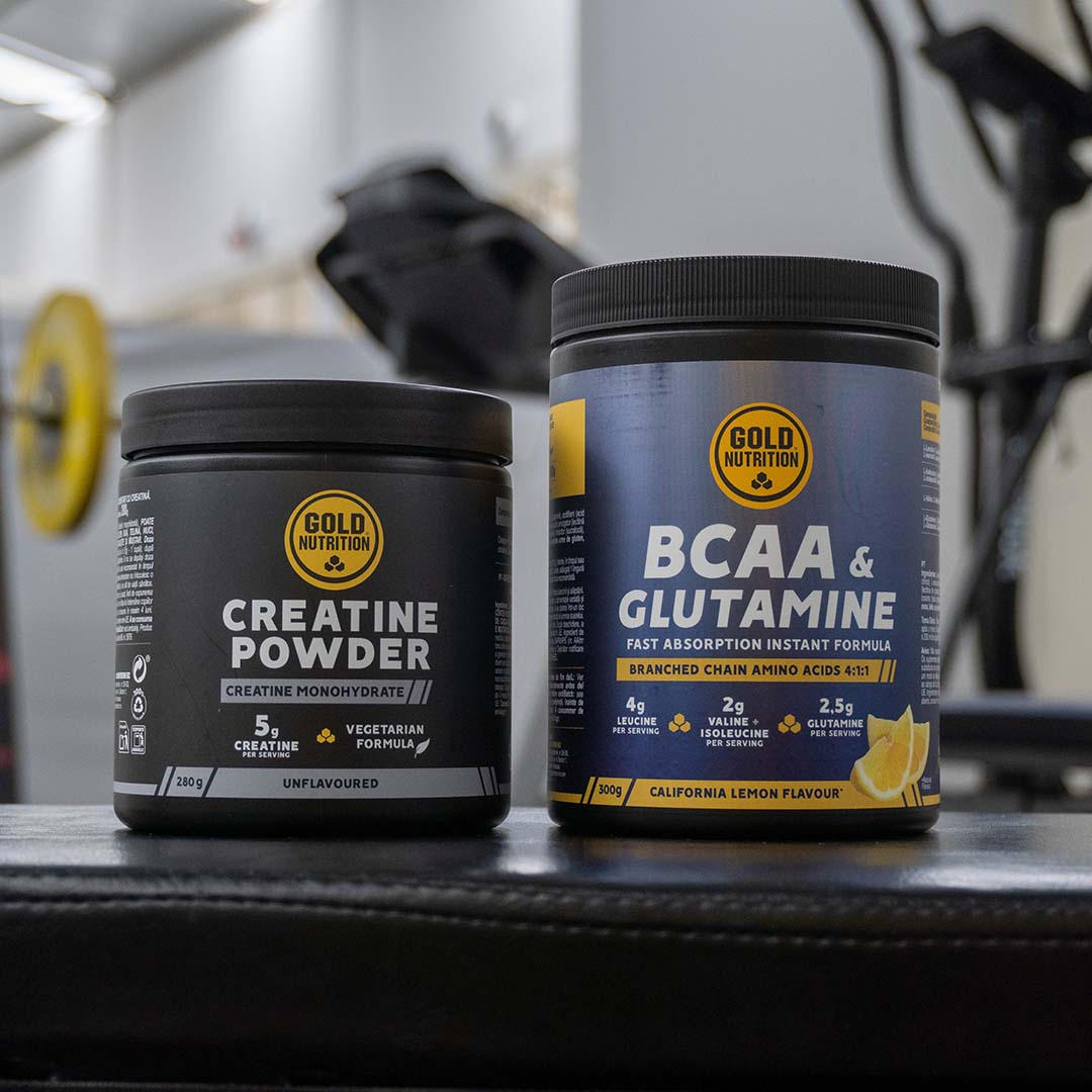 Dois suplementos da GoldNutrition sobre um banco de treino num ginásio. À esquerda, um frasco preto de Creatine Powder (Creatina Monohidratada) e, à direita, um frasco azul de BCAA & Glutamine (Aminoácidos de Cadeia Ramificada com Glutamina).