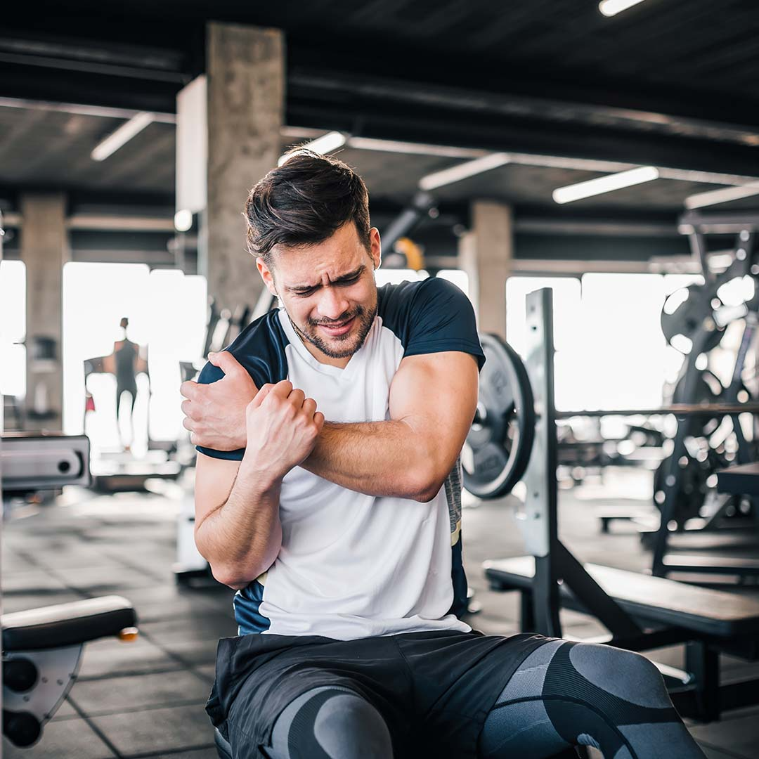 Homem jovem com expressão de dor segurando o braço no ginásio. Ele veste uma t-shirt branca e preta e calções pretos, sentado num banco de musculação com equipamentos de treino ao fundo.